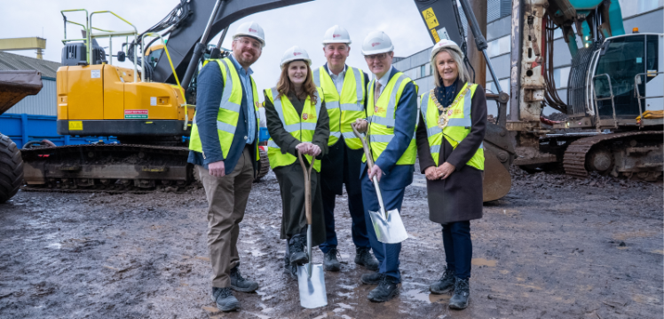A group of five people pose for the camera wearing high-vis vests and hard hats at a sod cut. Two of them are holding shovels.